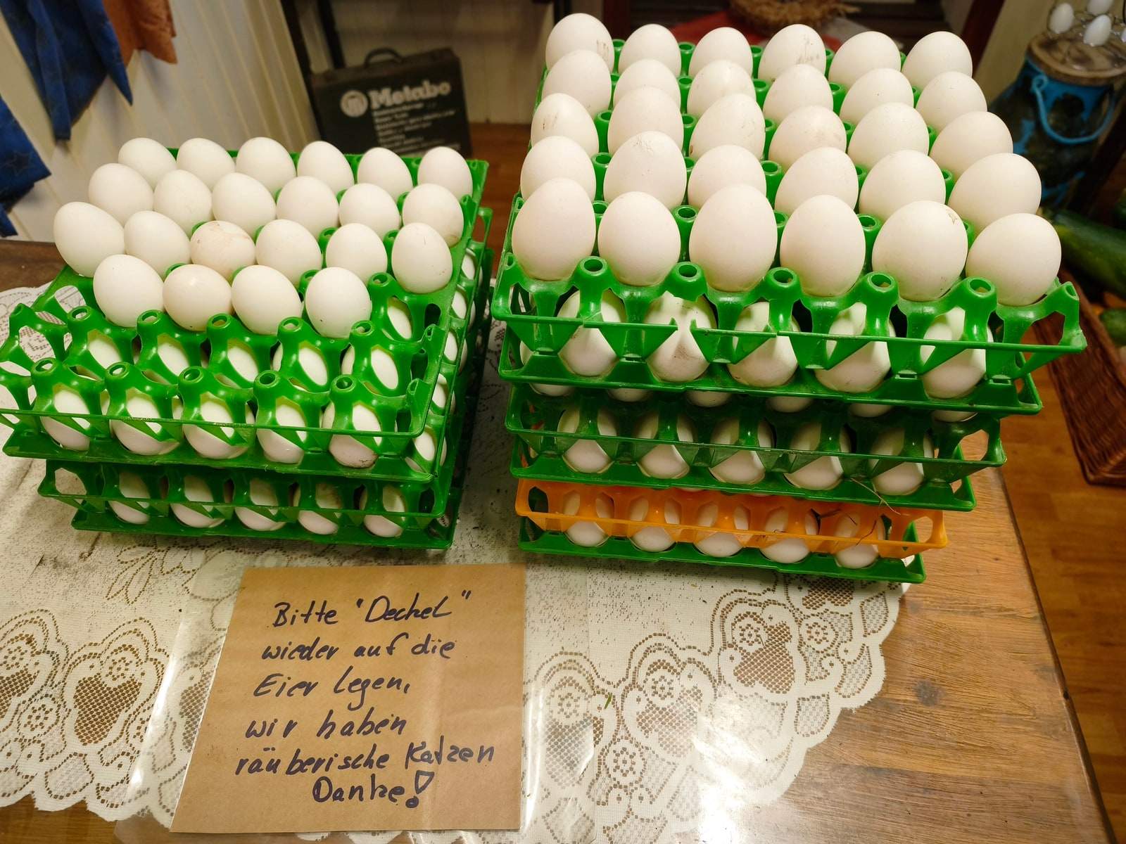 Zwei Stapel grüner Eierhorden voller weißer Eier stehen auf einem Holztisch mit Spitzendecke im Verkaufswagen; davor lehnt ein handgeschriebener Zettel mit der Bitte, den Deckel wieder auf die Eier zu legen.