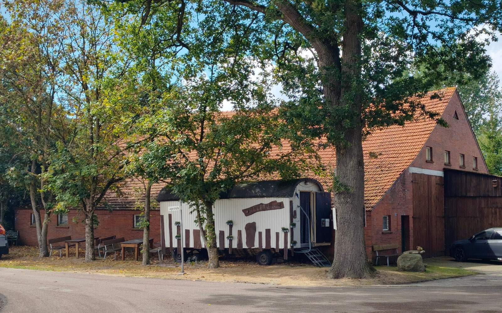 Der Hof von der Straßenecke aus: hinter alten Eichen steht der weiße Verkaufswagen an einer langen Klinkerscheune mit rotem Ziegeldach, links darunter zwei Tische mit Bänken.