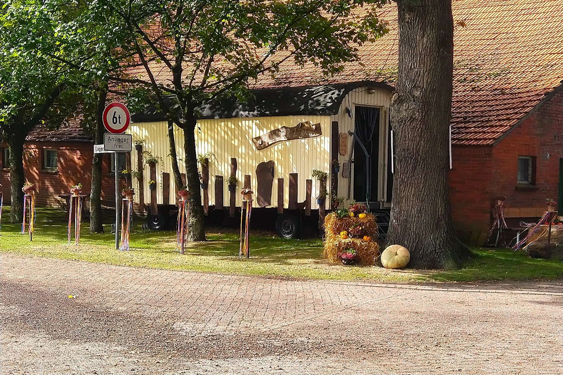 Der weiße Verkaufswagen steht der Länge nach unter einer alten Eiche vor dem roten Klinkerstall. Davor liegen Strohballen und ein heller Kürbis.